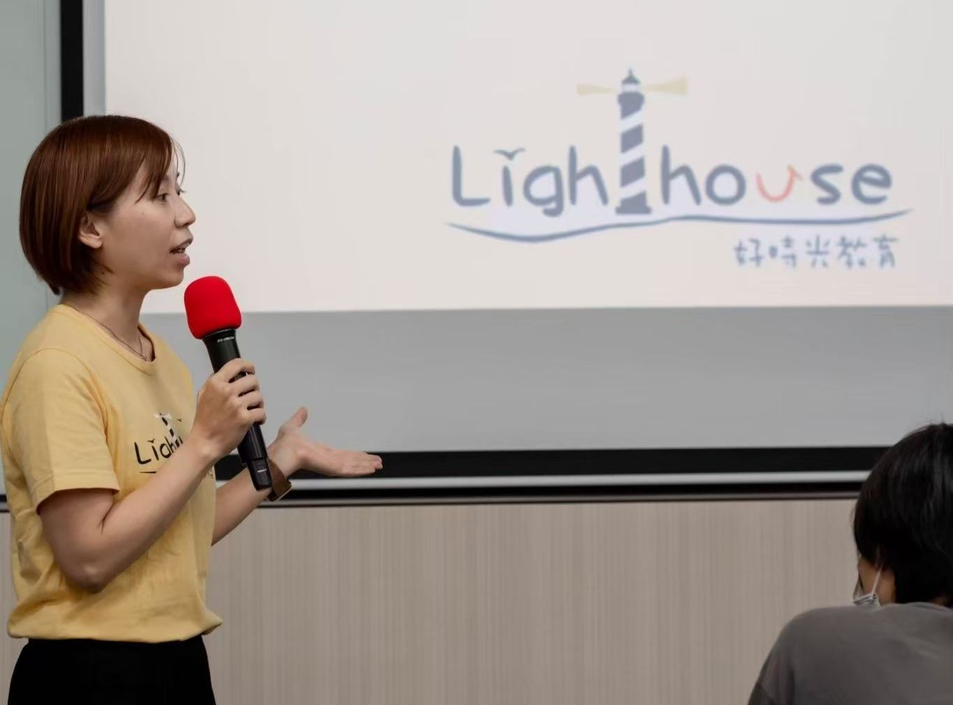 一位女士在「燈塔」活動上發表演講,她手持麥克風,背景中可以看到燈塔的標誌。