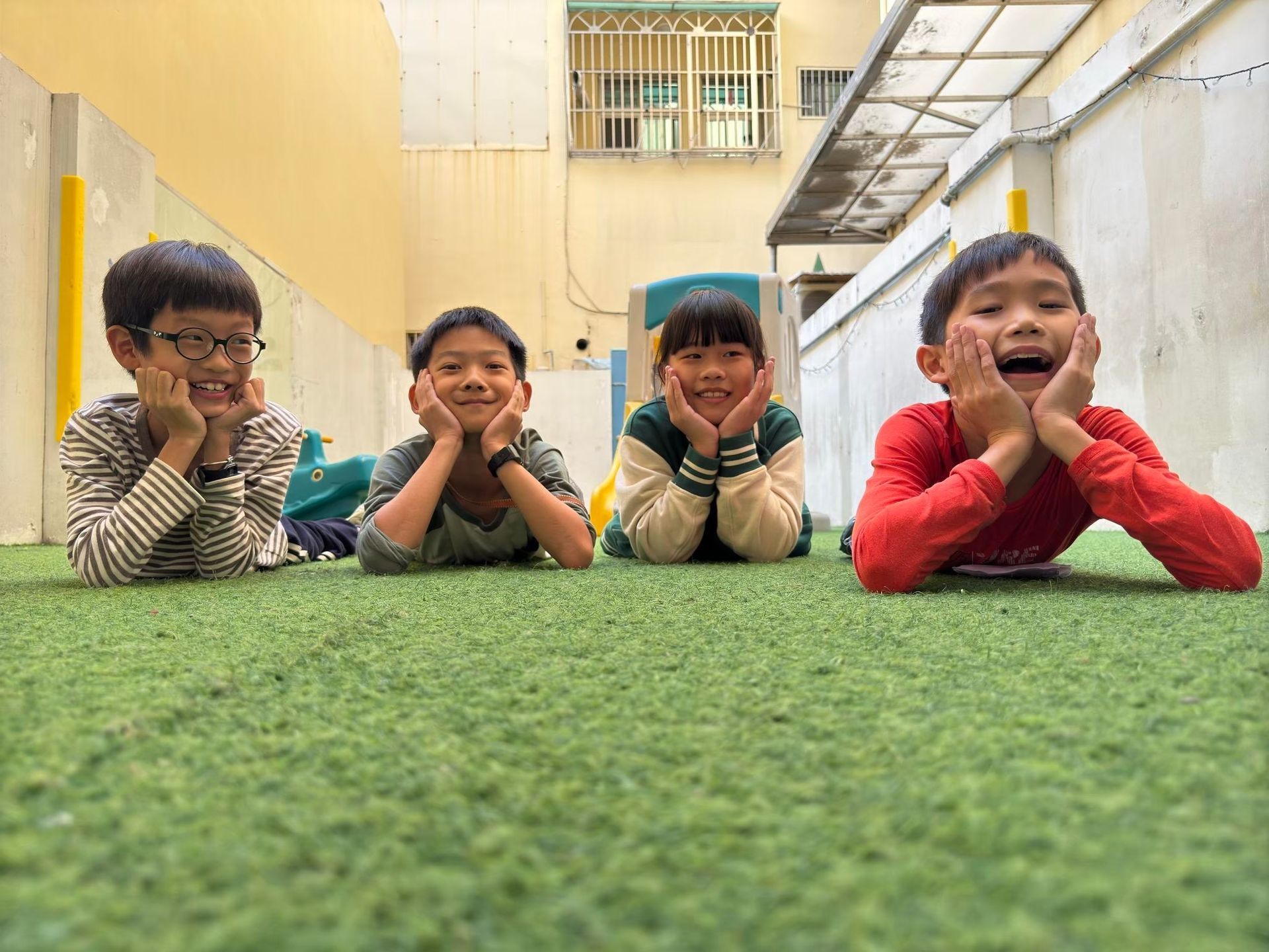 四個孩子麵帶微笑，躺在綠色的草坪上，雙手托著臉頰。淺色背景，戶外場景。