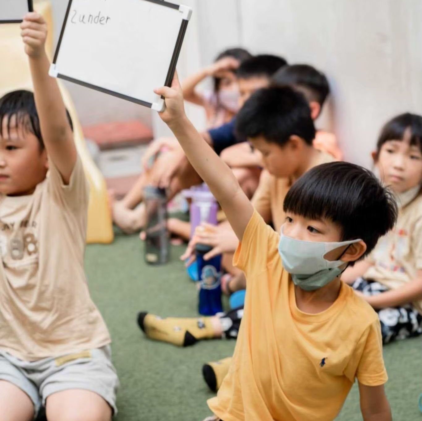 教室裡的孩子們舉起了手。其中一個孩子拿著一塊白板。有些孩子戴著口罩。