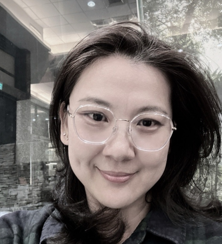 一位戴著圓框眼鏡的女子在建築物外微笑,臉頰露出酒窩。