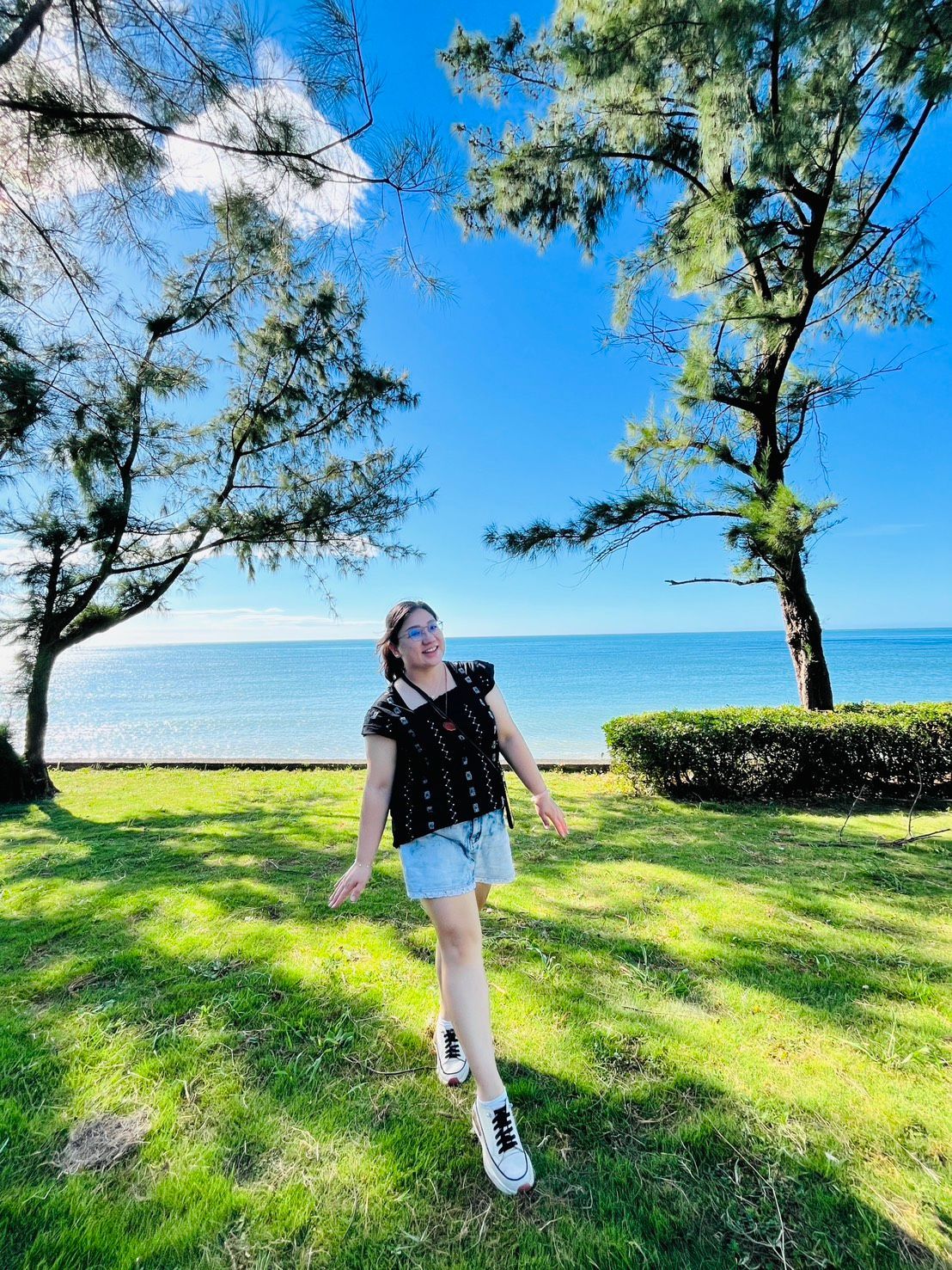 女人面帶微笑,站在水邊的草地上,樹木環繞著湛藍的天空。