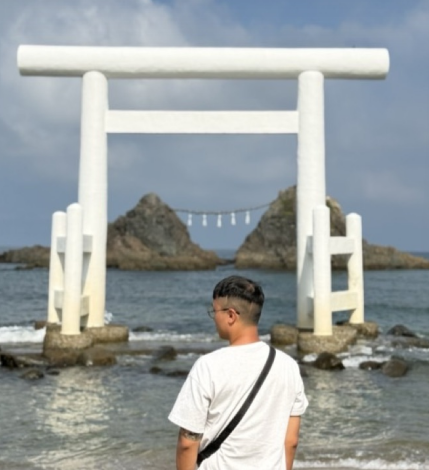 男子麵對淺水中的白色鳥居,背景是兩處岩石結構。