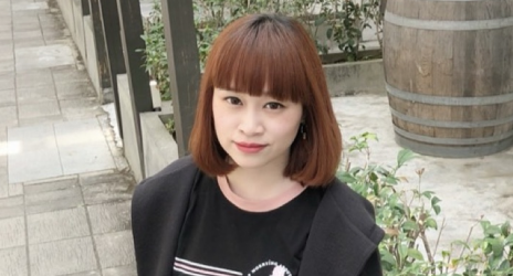 一位留著紅棕色短髮和瀏海的女子,身穿黑色襯衫和外套,正看向鏡頭。拍攝地點為戶外。