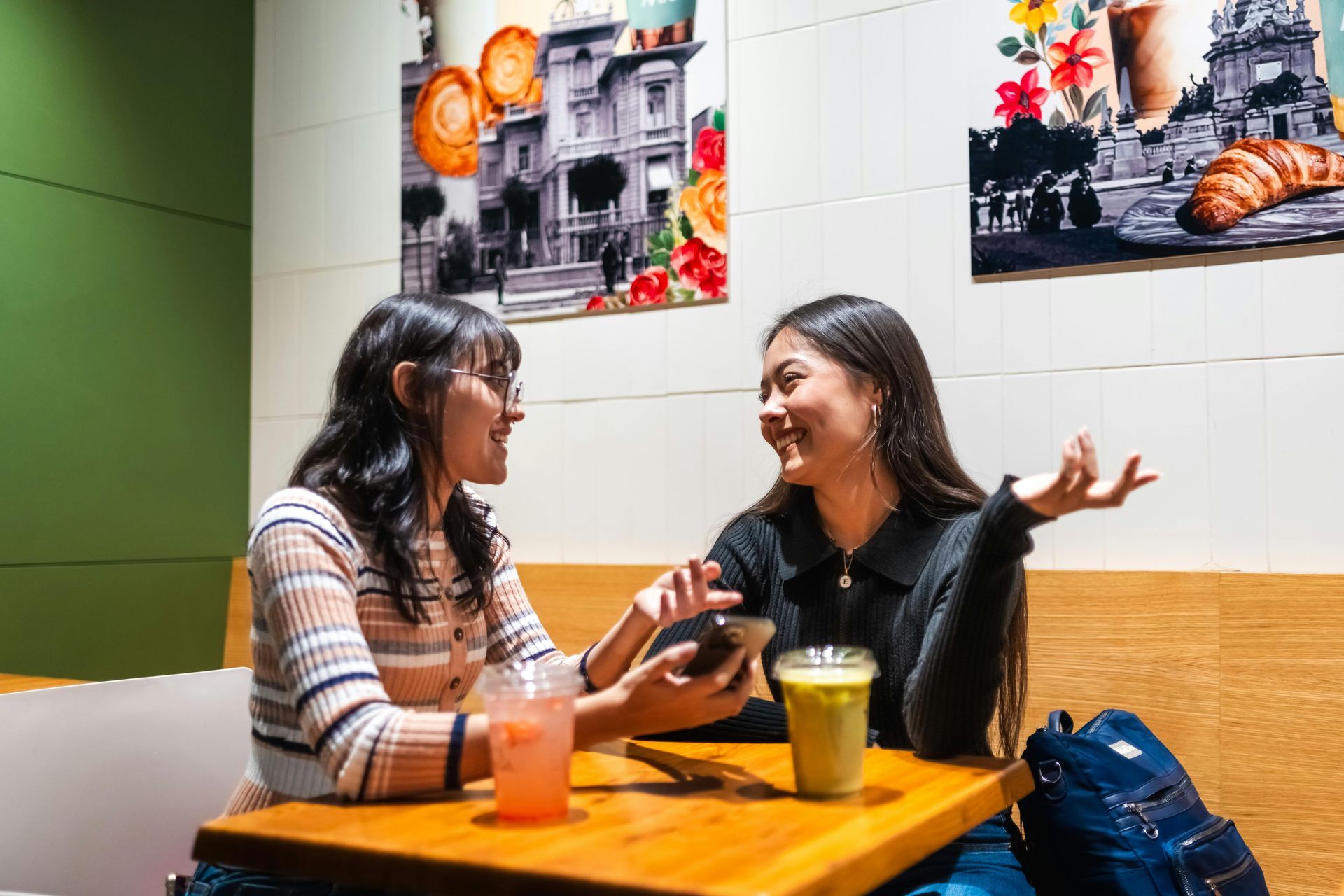 兩名女子麵帶微笑,坐在咖啡館裡的一張桌子旁,桌上放著飲料,她們正在看手機。