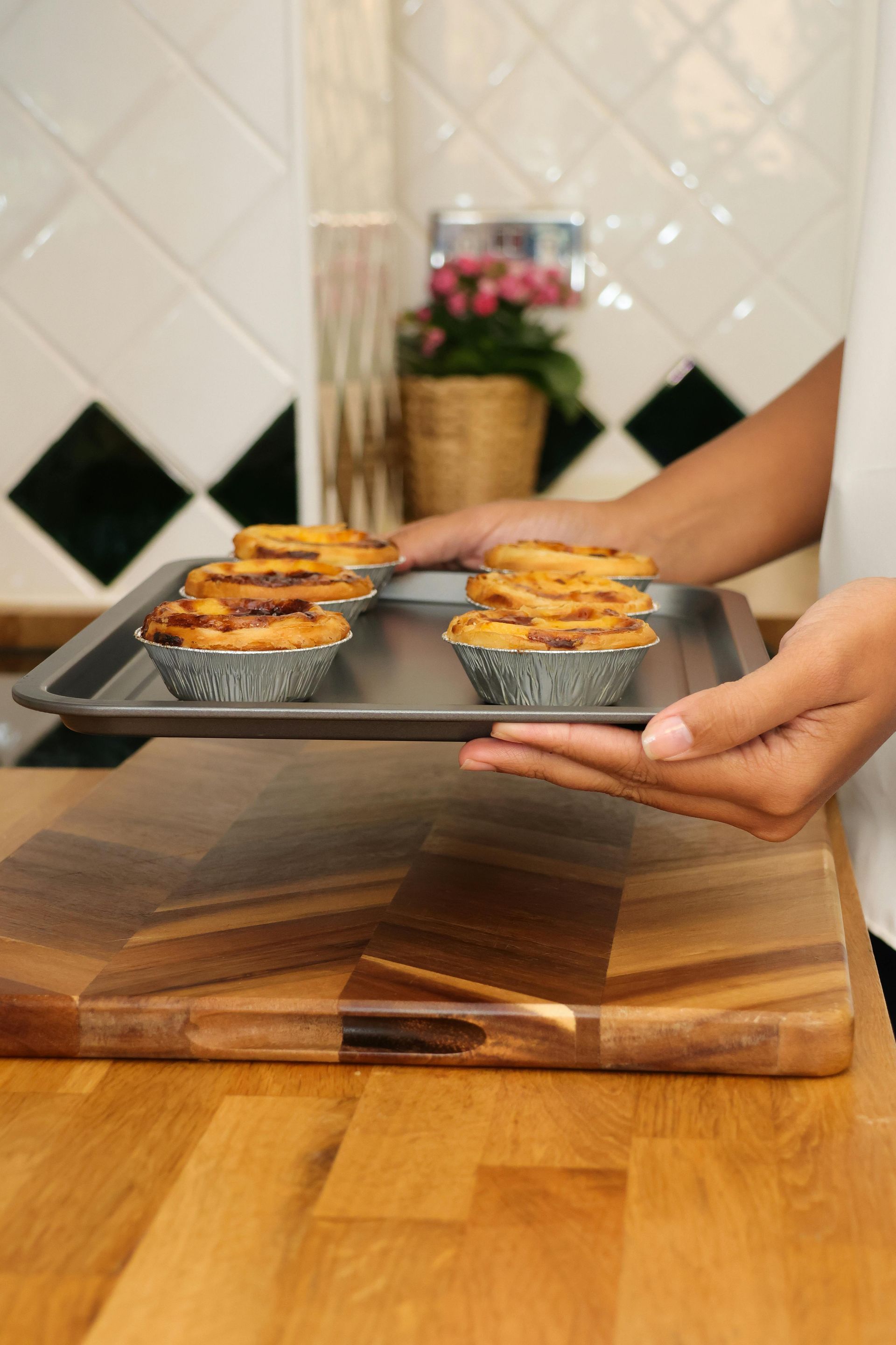 一個人手捧一盤烤好的蛋塔，廚房的防濺板貼有磁磚，檯面是木質的。