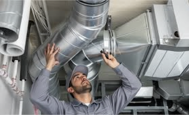 HVAC technician inspecting ventilation ducts in a building. He's wearing a gray uniform and a cap.