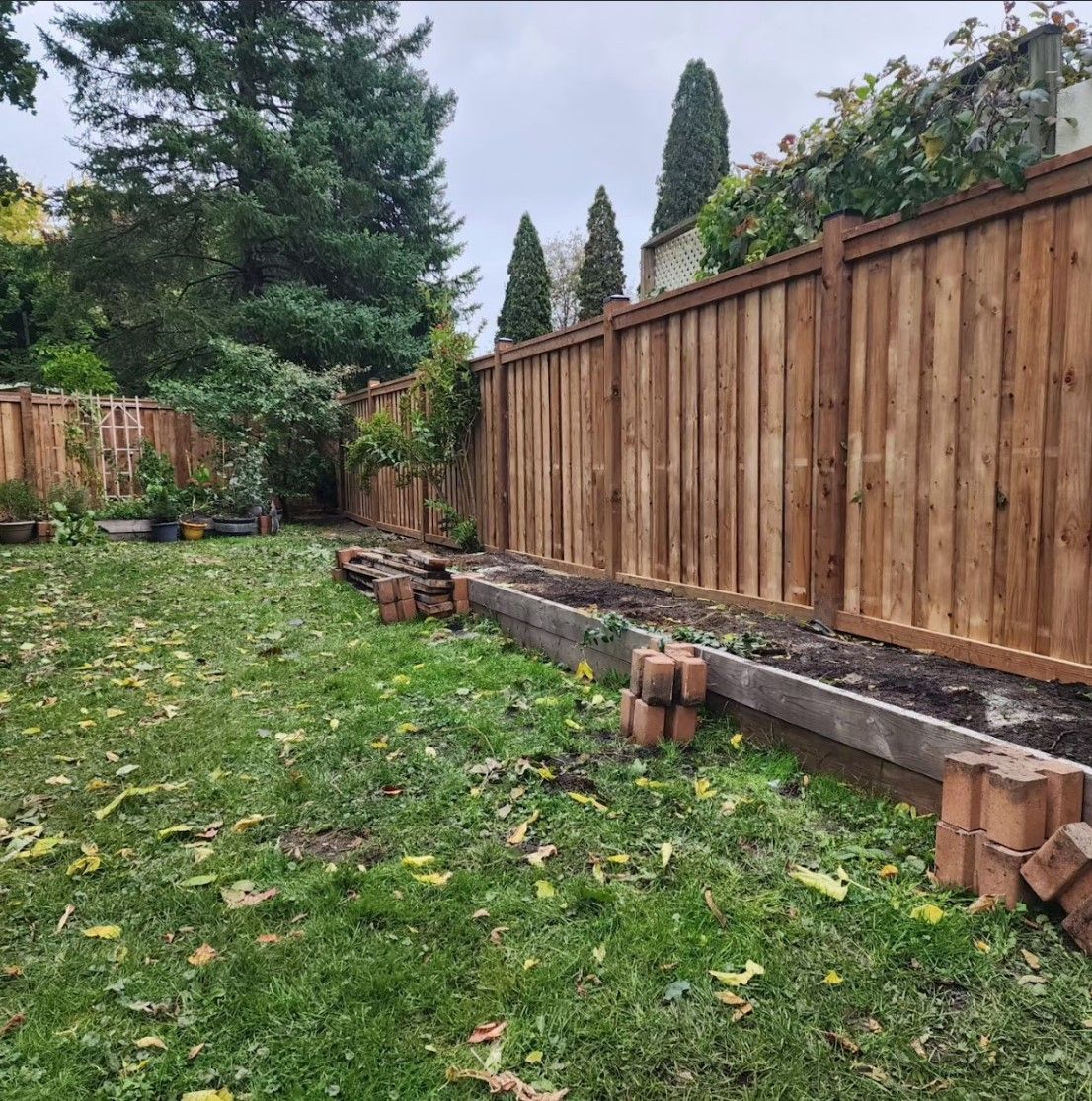 Wooden fence bordering a grassy backyard with raised garden beds and brick edging. Overcast sky.