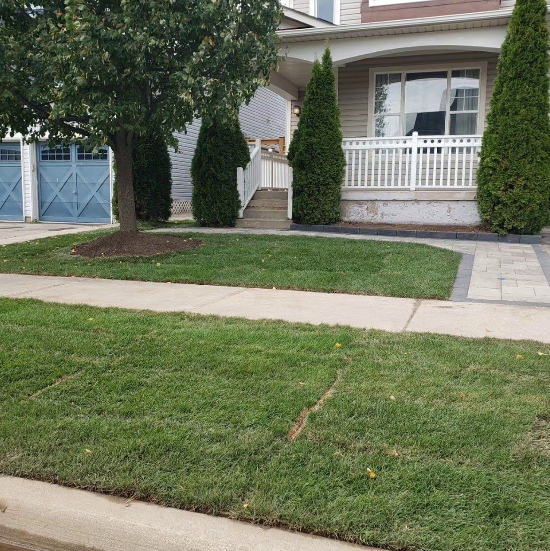 Lush green lawn in front of a house with a white porch and blue garage doors.