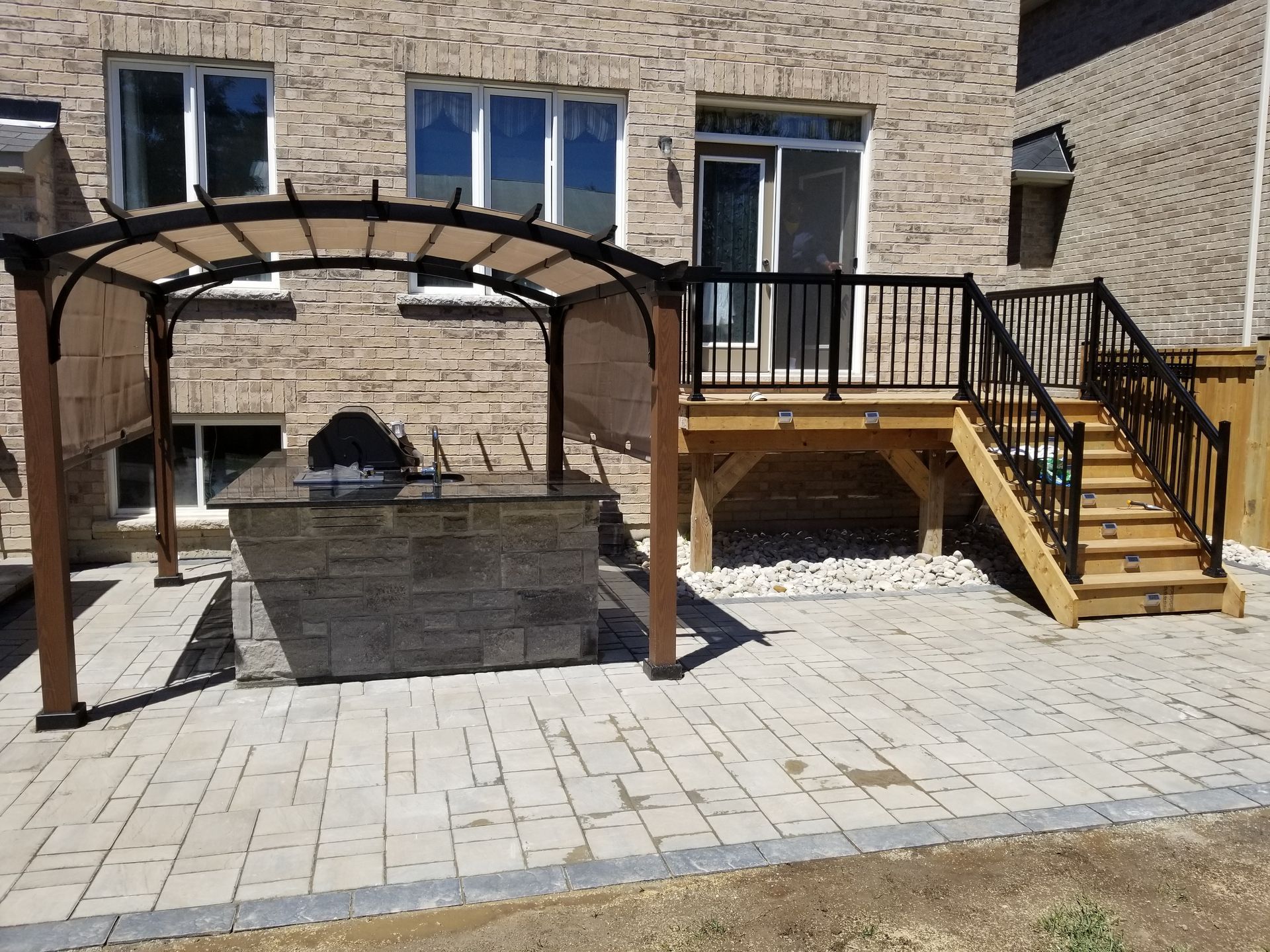 Backyard patio with built-in grill, pergola, and elevated wooden deck with black railing.