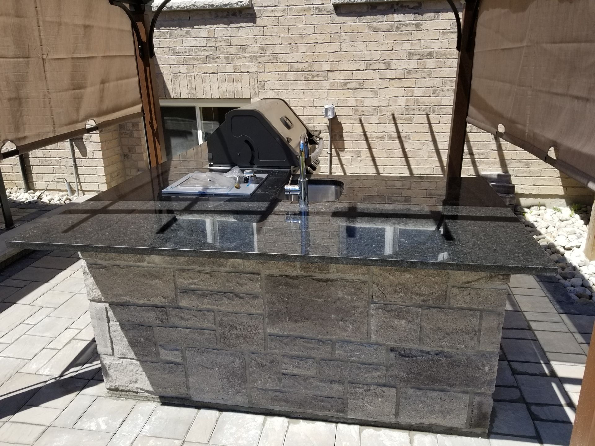 Outdoor stone-faced kitchen island with a grill and sink, topped with black granite.  Beneath a shaded pergola.
