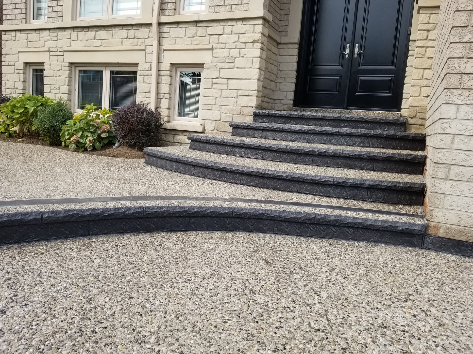 Exterior view of a house with concrete steps leading to black double doors. Walkway and steps have dark edging.