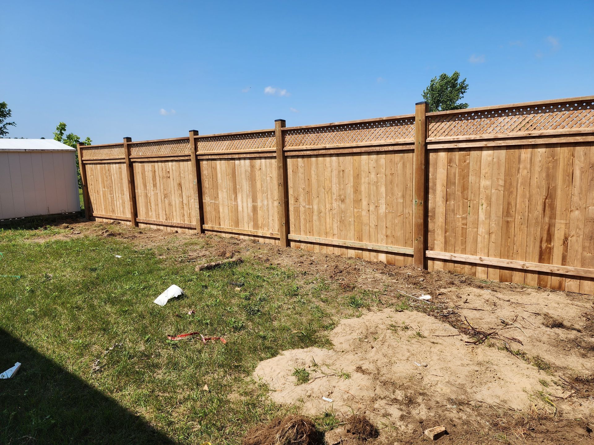 Wooden fence in a backyard with patchy grass and a pile of sand under a clear blue sky.
