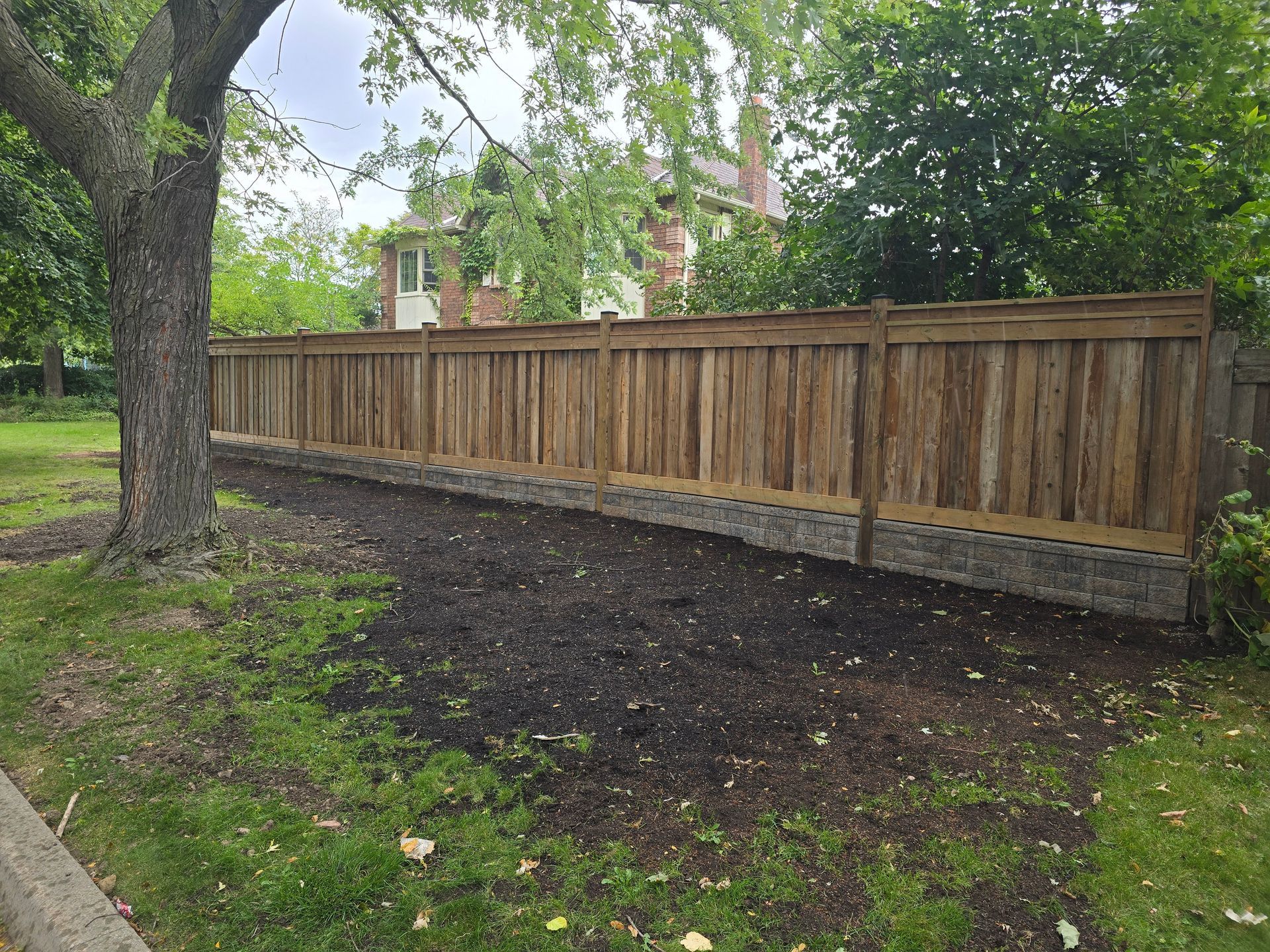 Wooden fence bordering a yard with fresh mulch and grass, trees in the background.