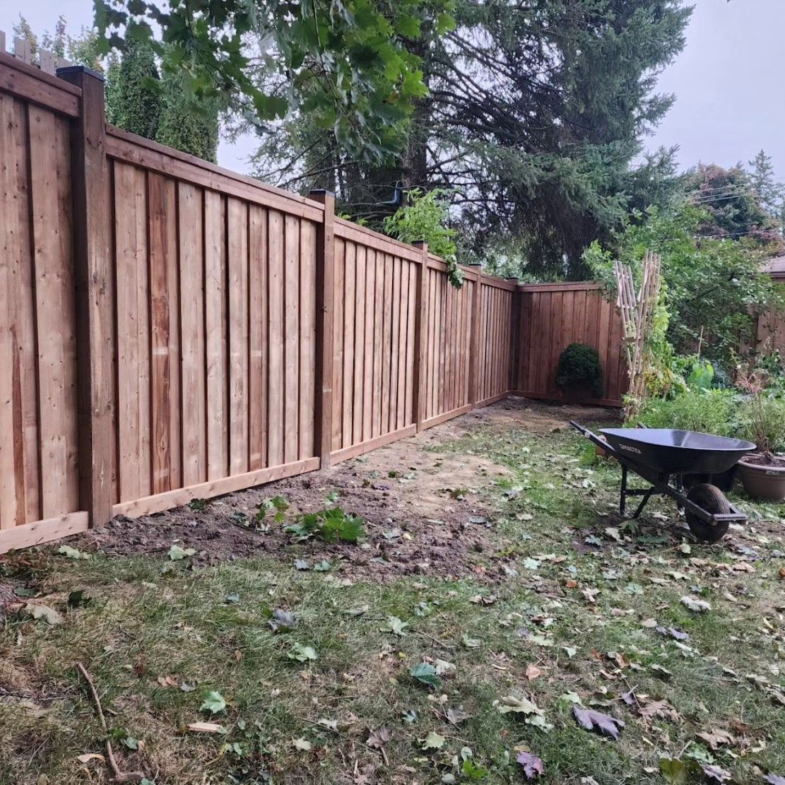 Wooden fence bordering a backyard with a wheelbarrow. Brown wood, green grass, overcast.