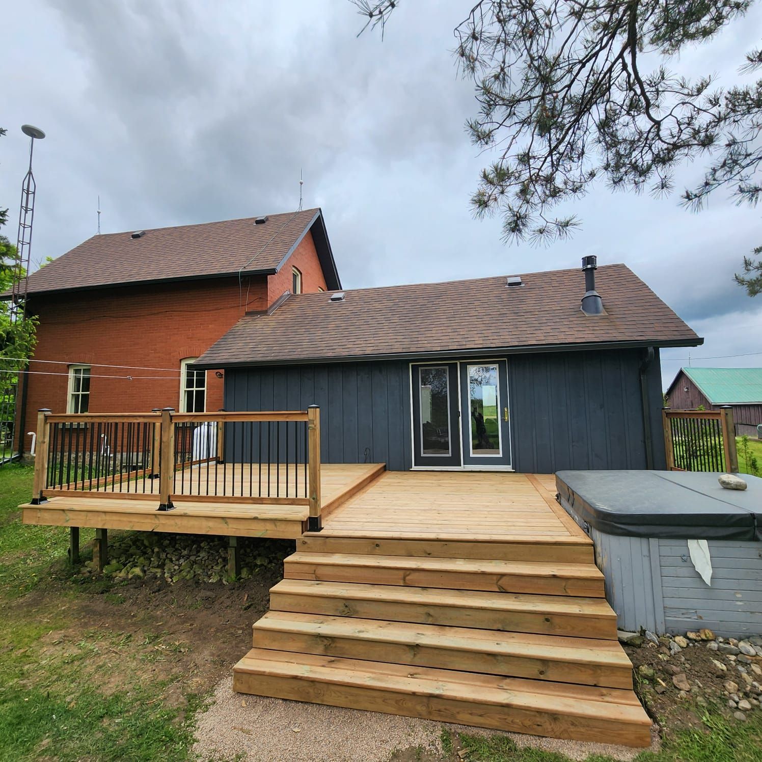 Wooden deck with stairs, attached to a dark blue house. A red house is behind it. Hot tub on the deck.