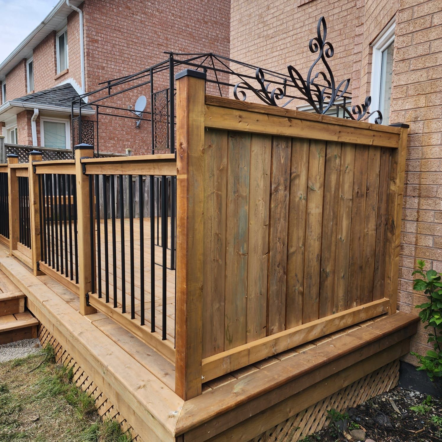 Wooden deck with a stained fence and black metal railing, near a brick building.