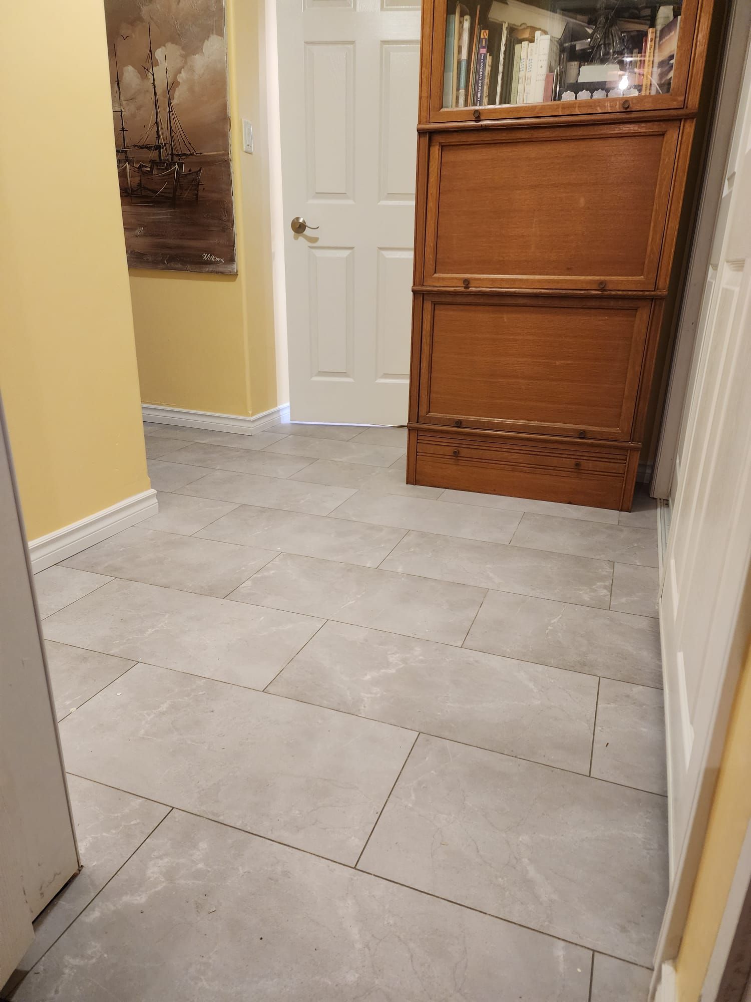 Hallway with gray tiled floor, a white door, and a wooden cabinet. Light yellow walls.