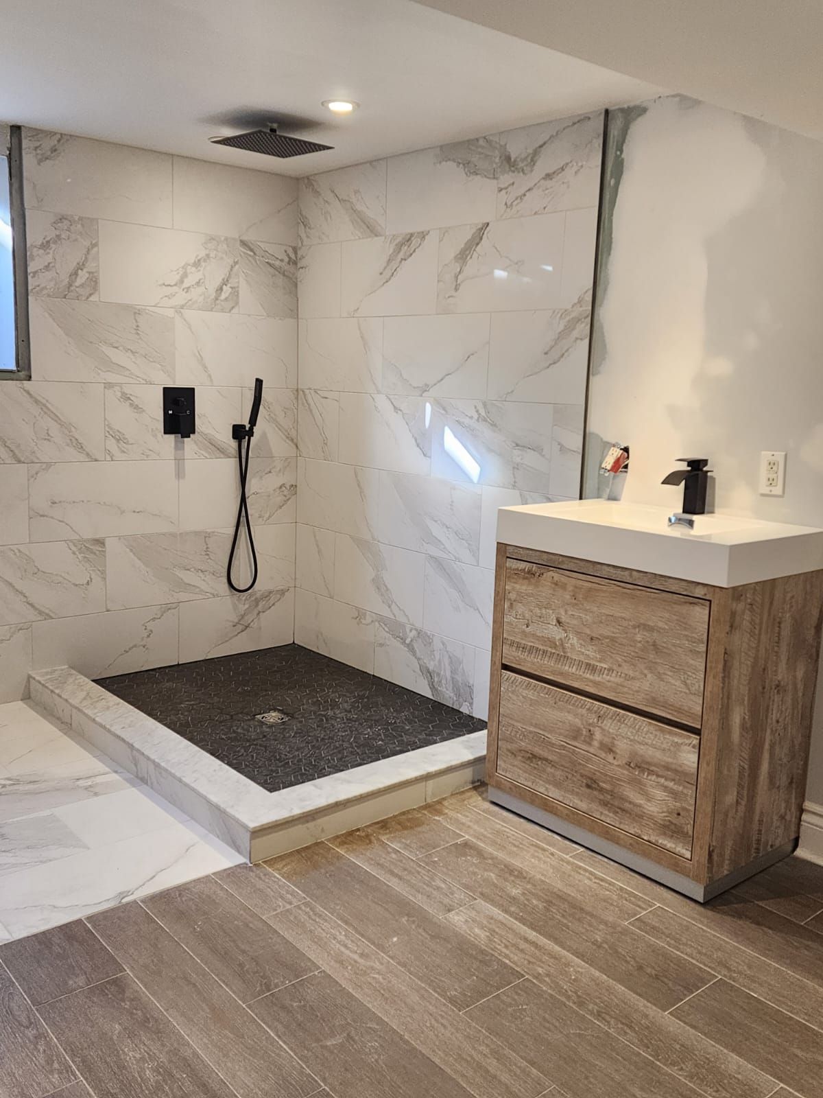 Modern bathroom with marble tile shower, black fixtures, wooden vanity, and hardwood floor.