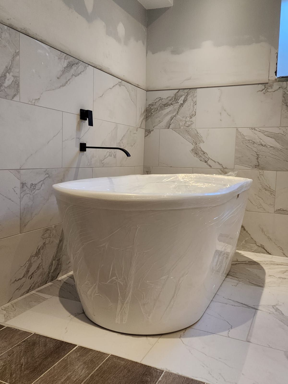 Freestanding white bathtub with black faucet, marble tile walls, and a window in a bathroom.