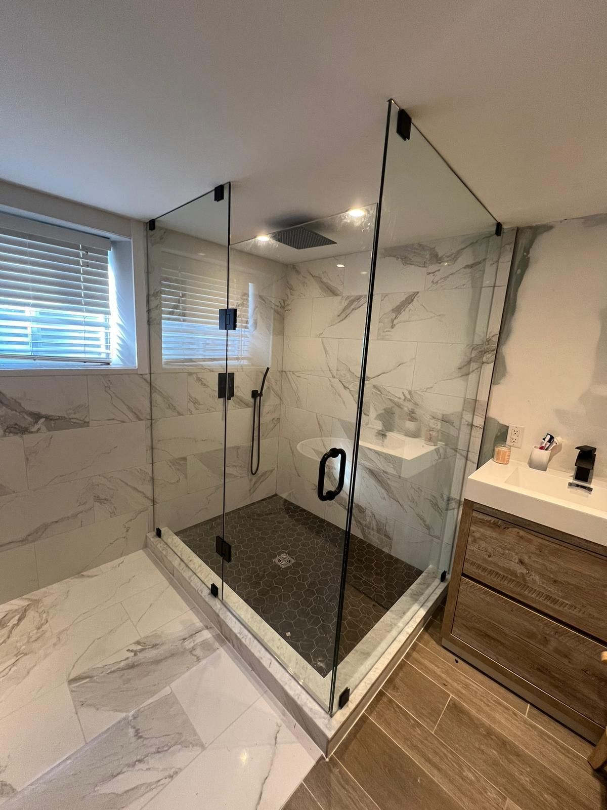 Modern glass shower enclosure in bathroom with marble walls and wood vanity.