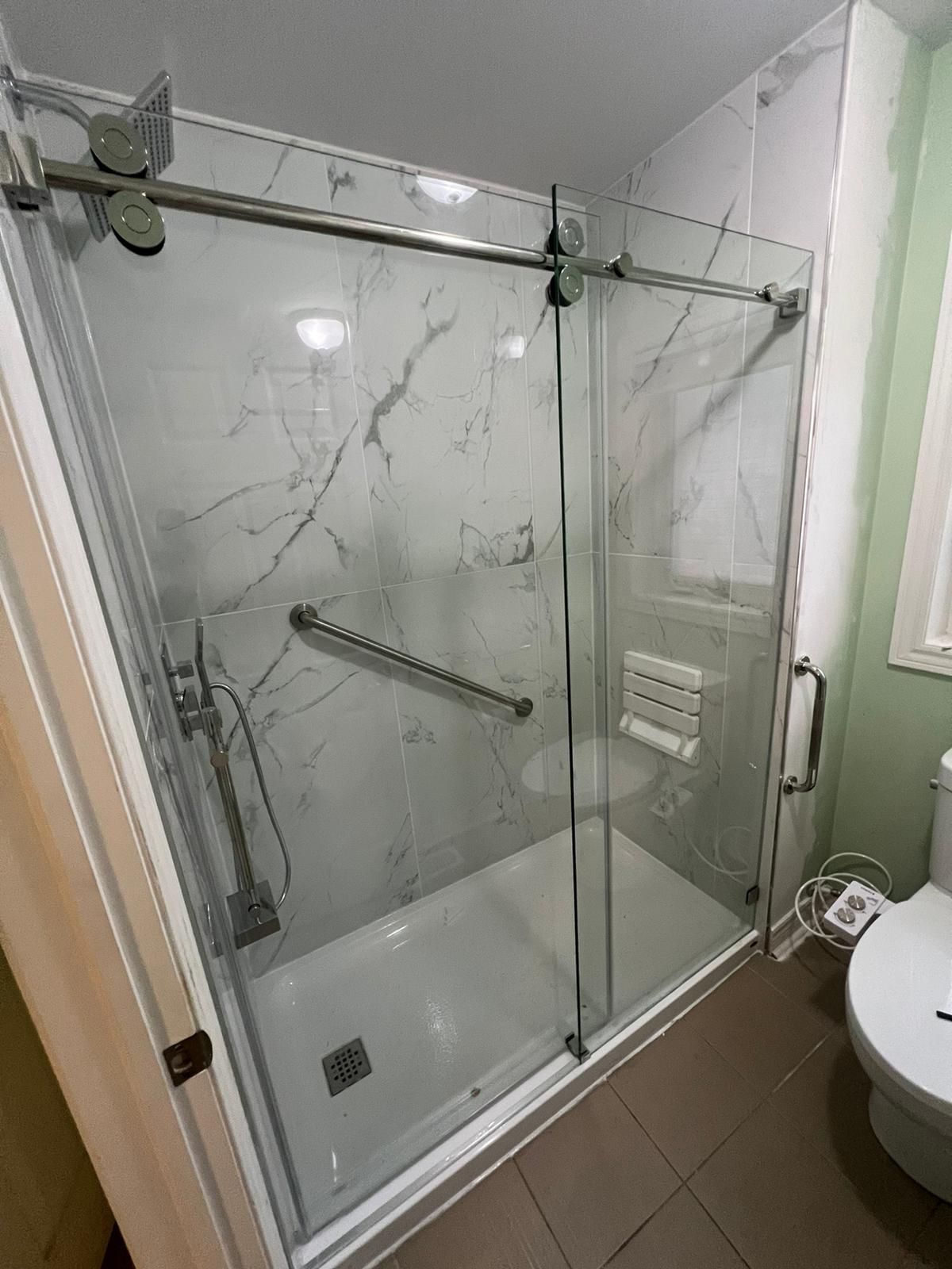 Shower stall with marble-patterned tile, sliding glass doors, and a grab bar in a bathroom with a toilet.