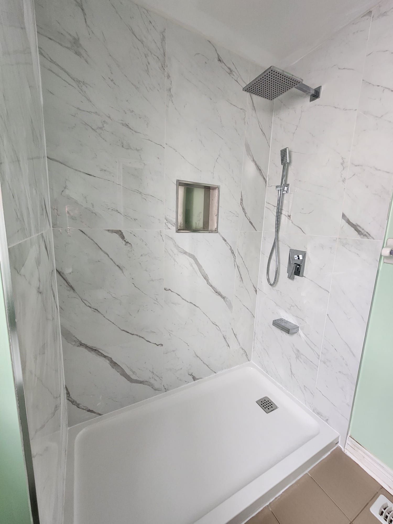 Shower stall with white marble-like tiles, silver shower head, and built-in soap dish.