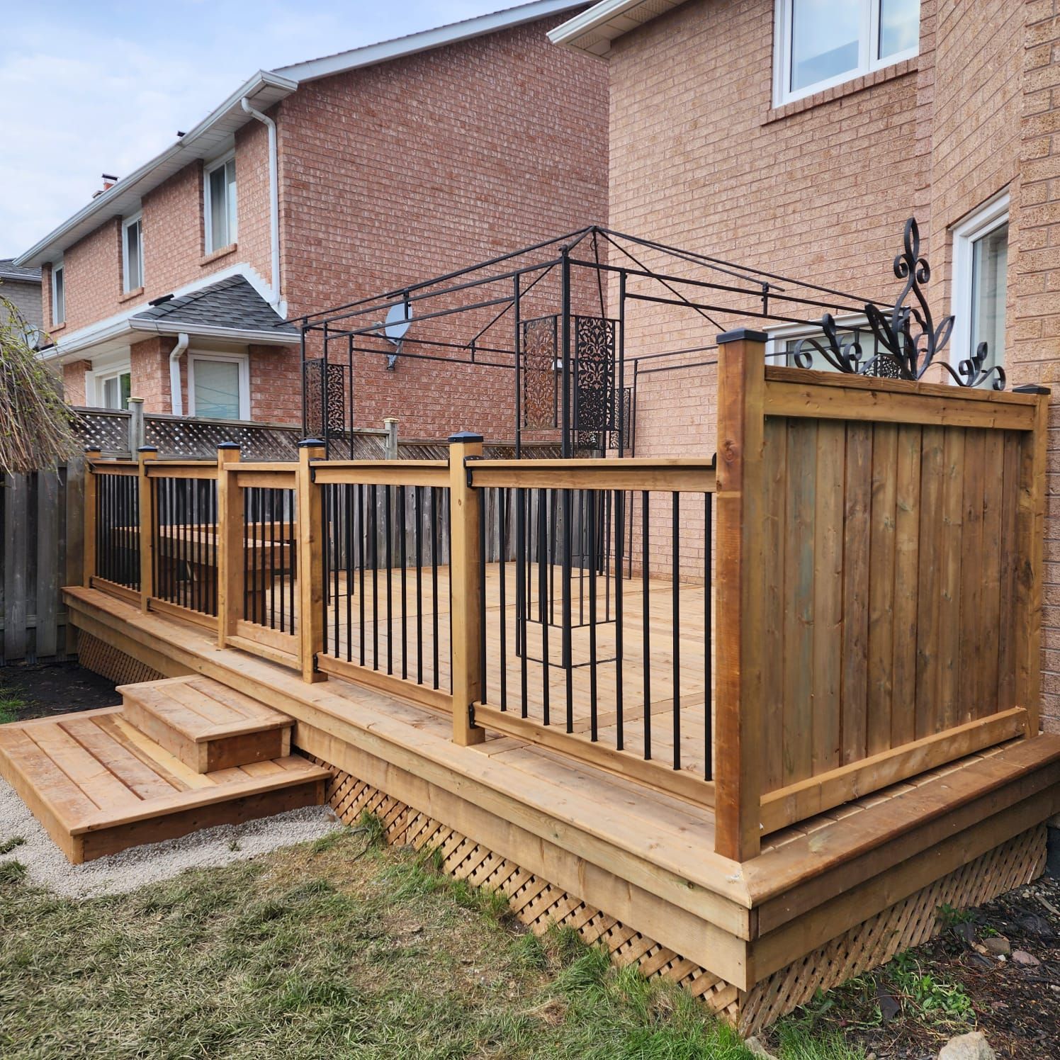 Wooden backyard deck with steps, railings, and gazebo frame, next to a brick building.