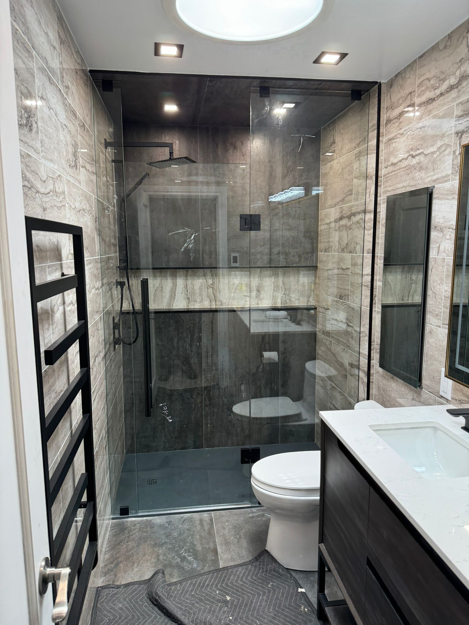 Modern bathroom with glass shower enclosure, gray stone-like tiles, toilet, and a black heated towel rack.
