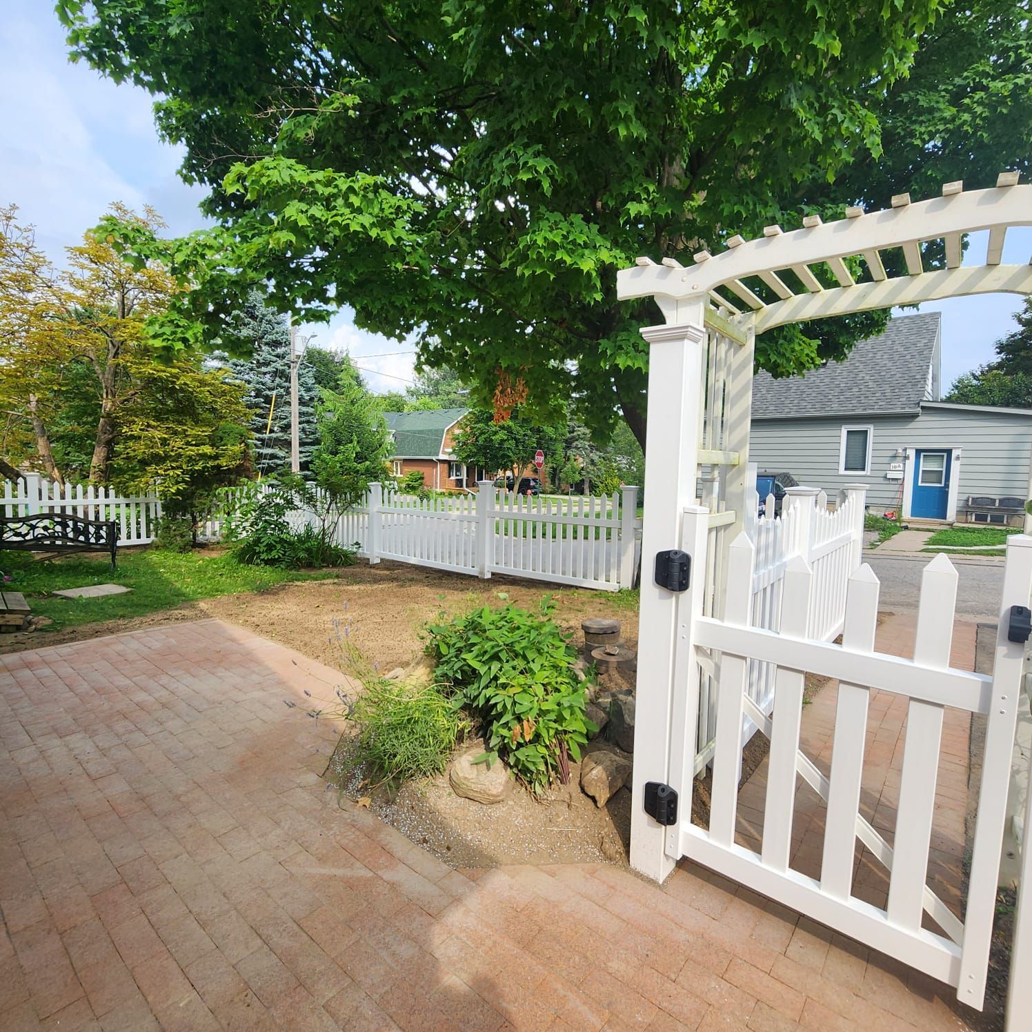 White picket fence with gate, brick patio, green trees, and small house in the background.