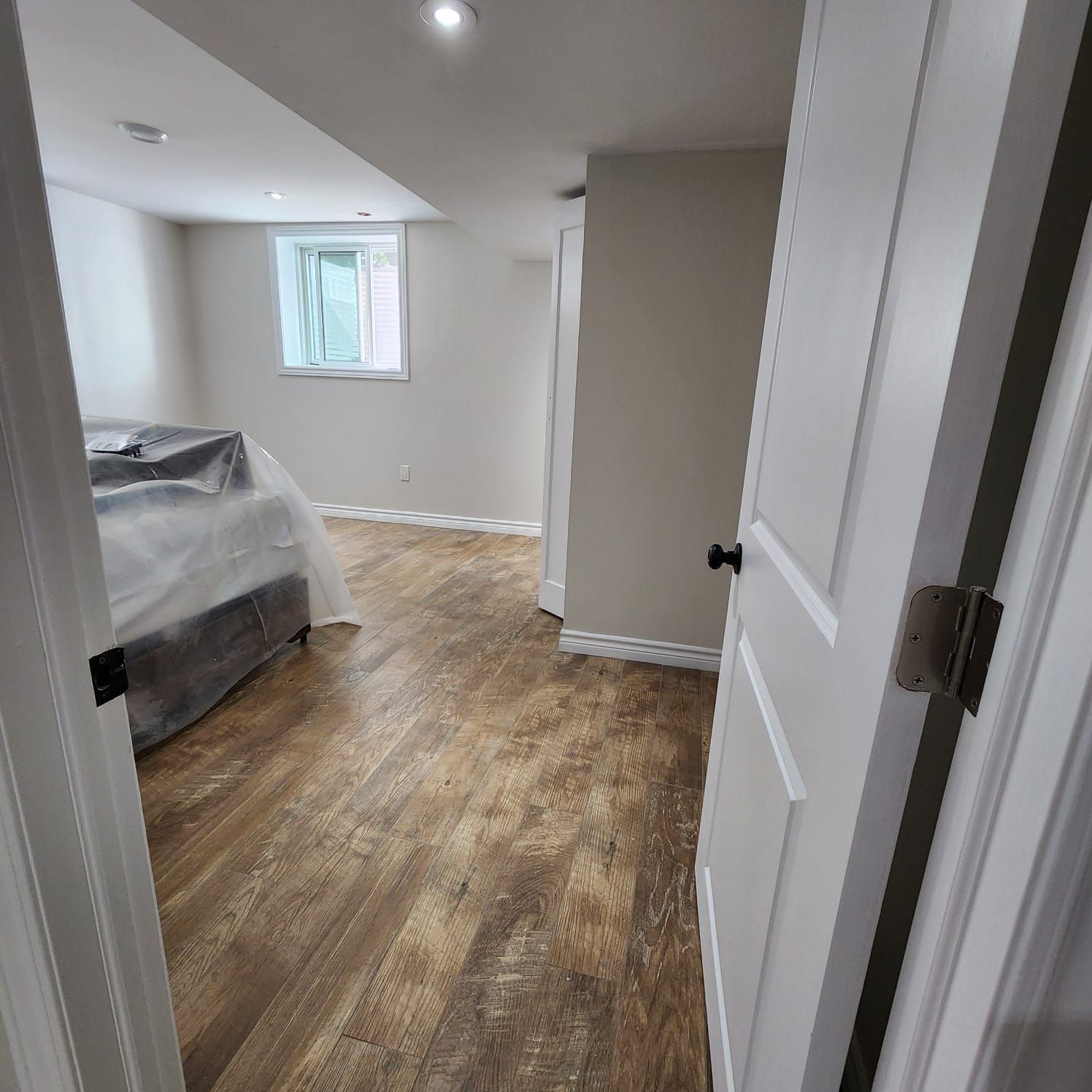 Open white door reveals a renovated basement with wood-look flooring and gray walls. A bed is covered in plastic.