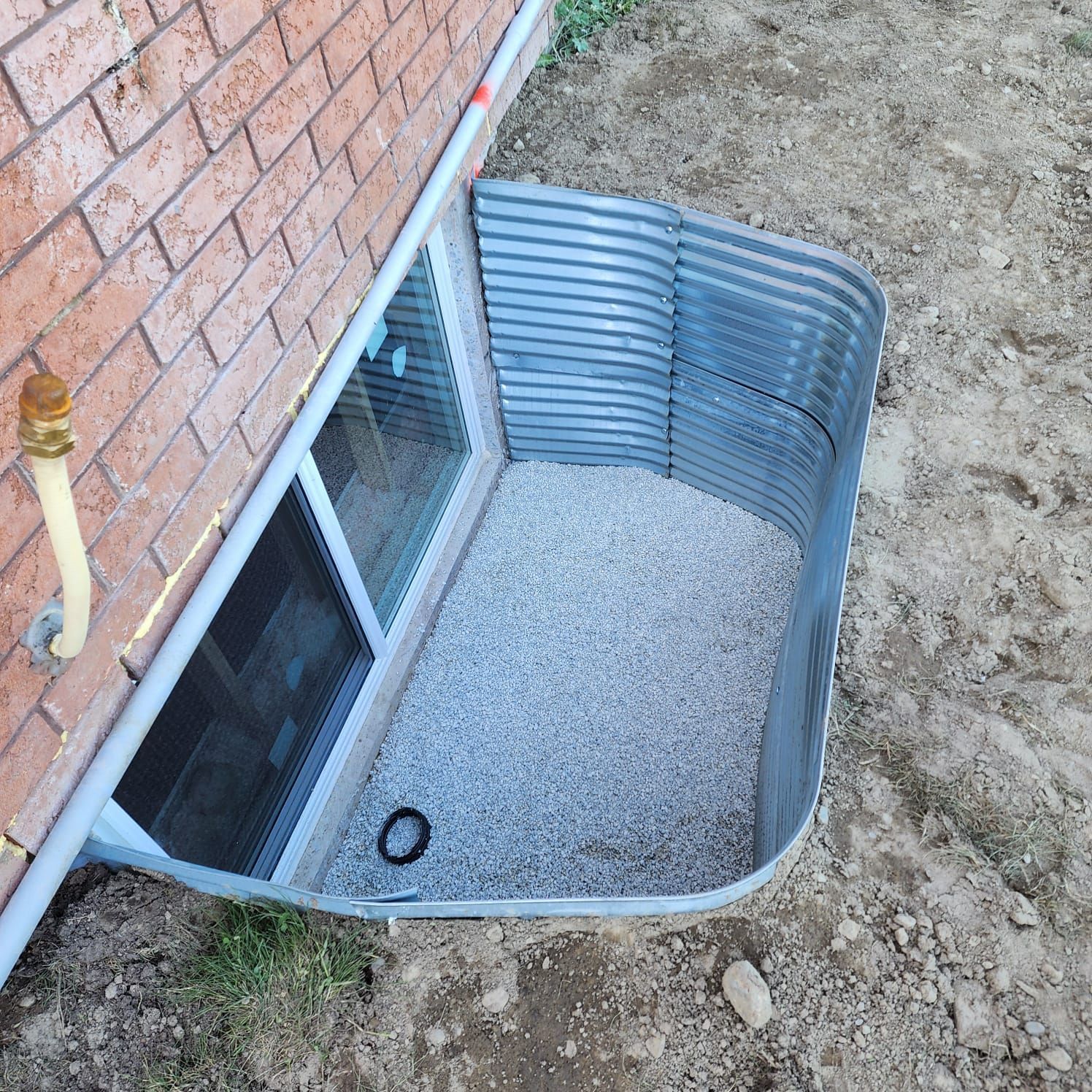 Window well with a metal liner, gravel base, and a window in a brick wall.