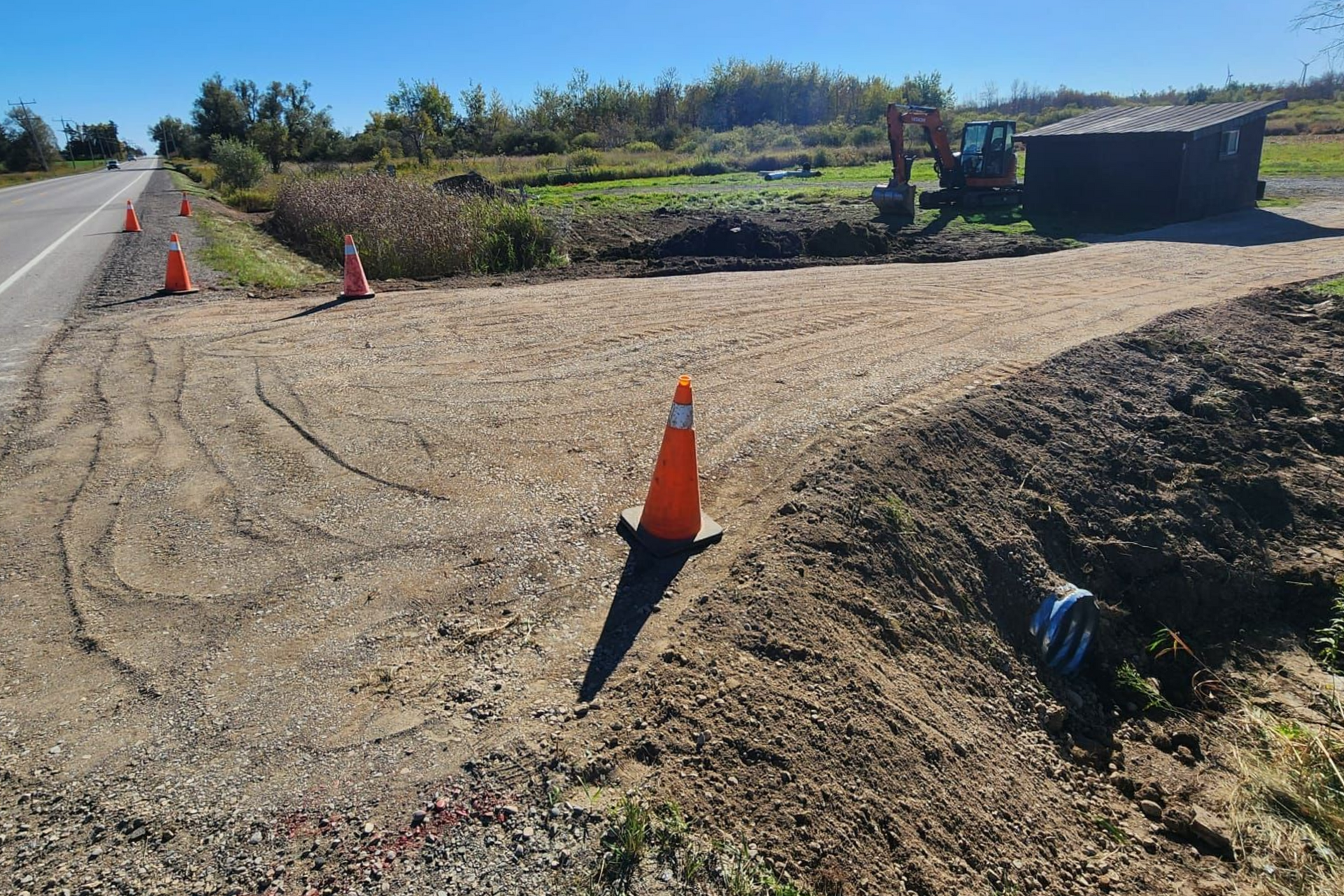 Roadside construction scene with orange cones, gravel, and an excavator. Blue sky and rural setting.