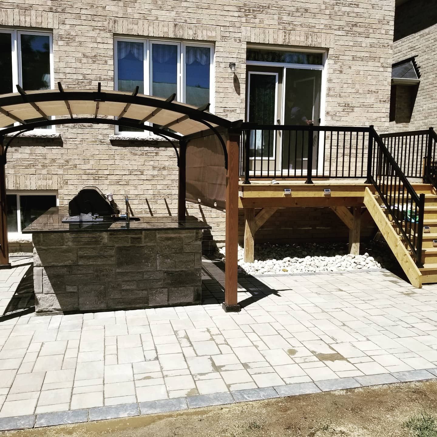 Backyard patio with built-in grill under a pergola, adjacent to a wooden deck with railings, and brick house.