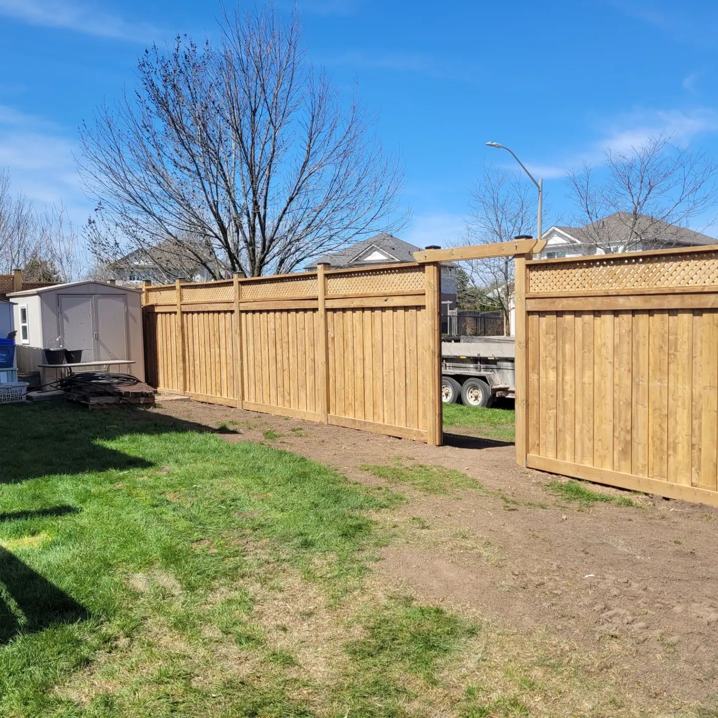 Wooden fence with open gate in a backyard. Light brown wood, green grass, blue sky.