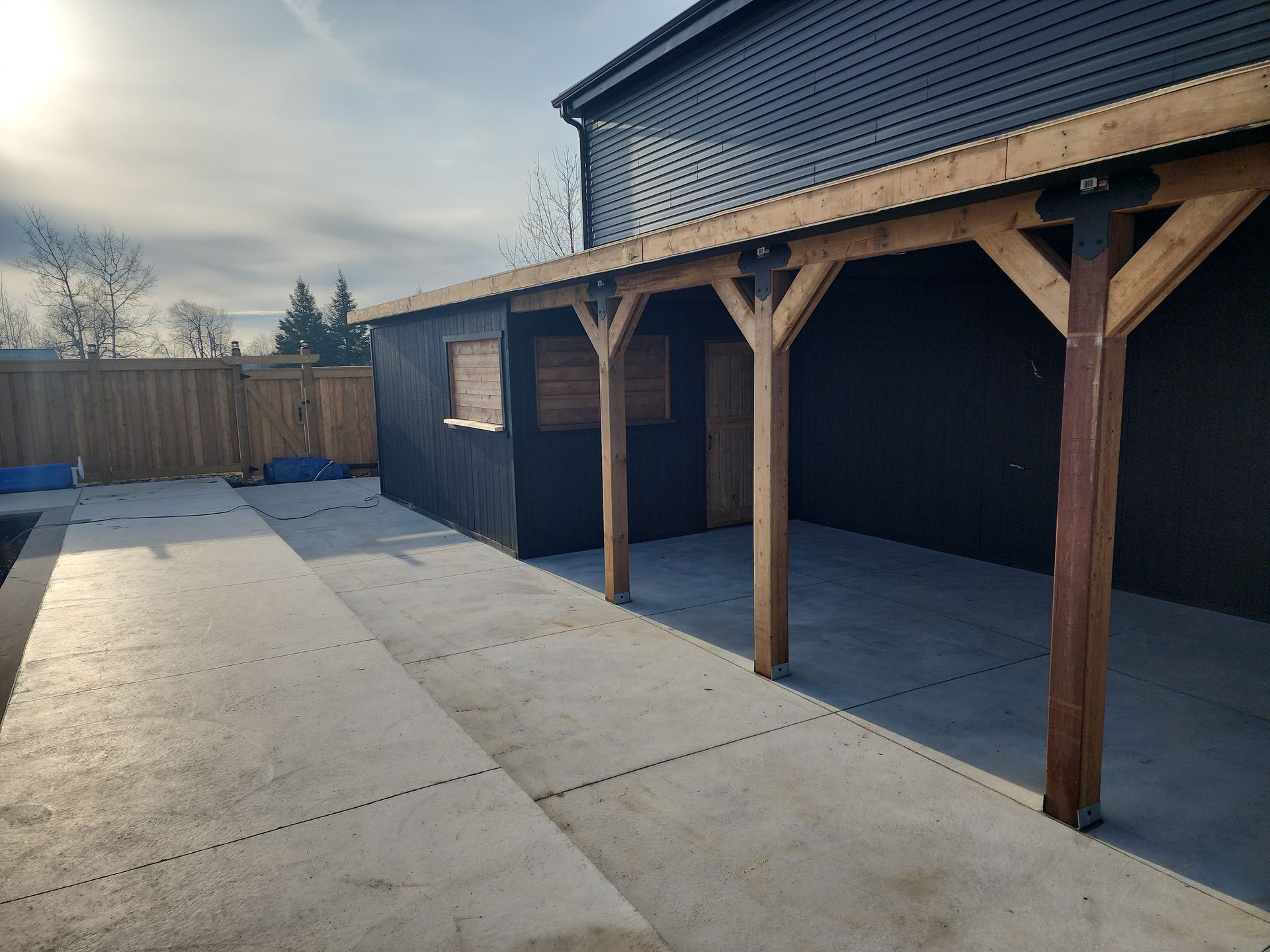 Concrete patio with a dark wooden shed and covered seating area under a sunny sky.