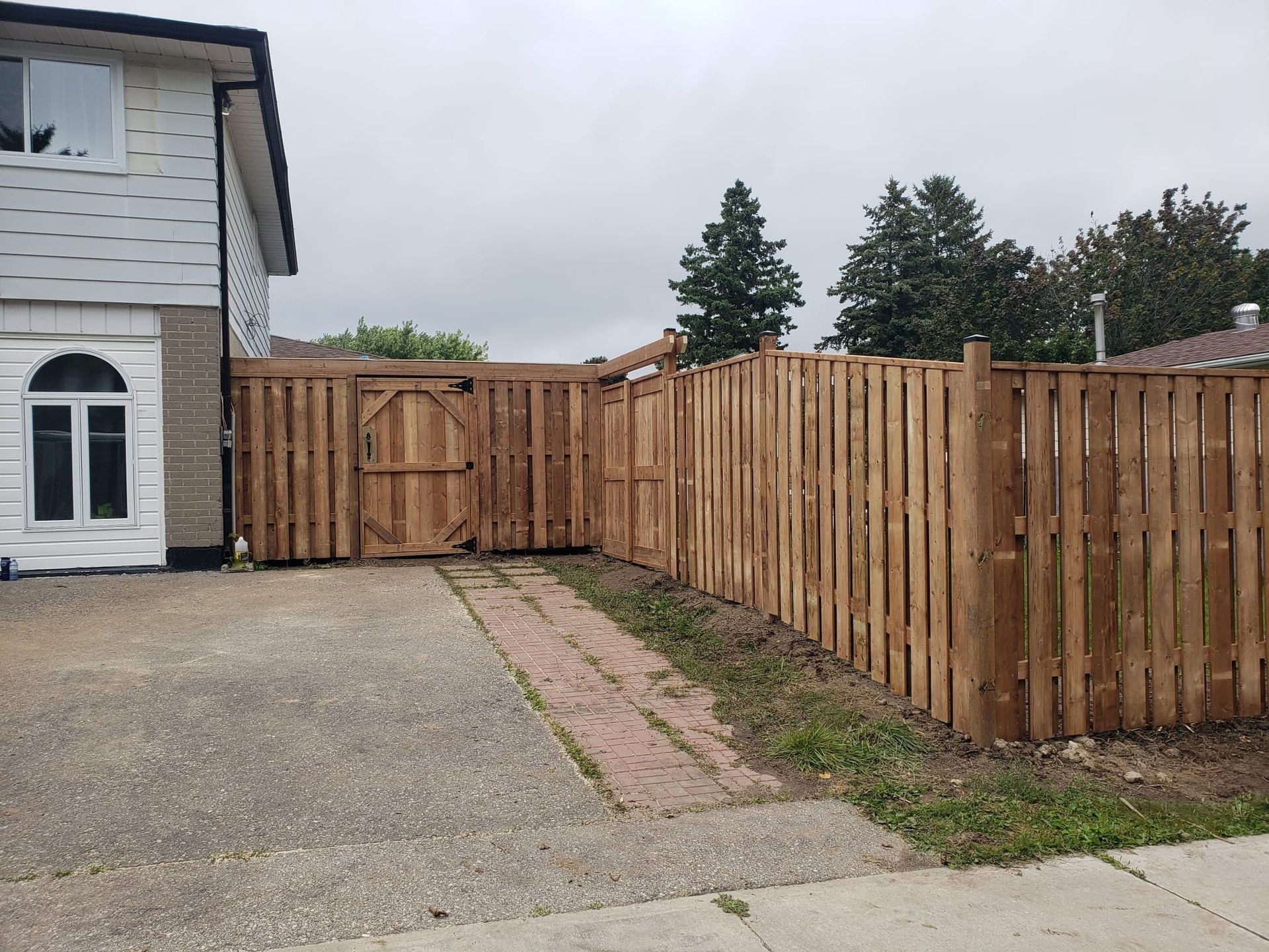 Brown wooden fence with a gate encloses a driveway next to a two-story house under a cloudy sky.