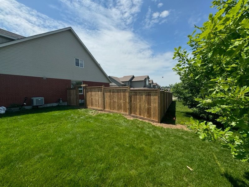 Wooden fence surrounds a backyard, next to a house with red brick and green grass under a blue sky.