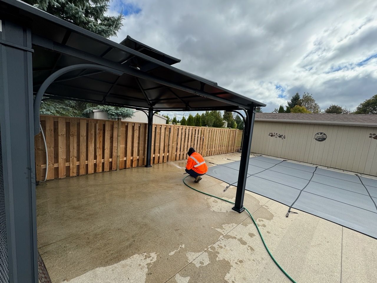 Person in orange jacket hosing down a concrete patio near a gazebo and a pool with a cover.