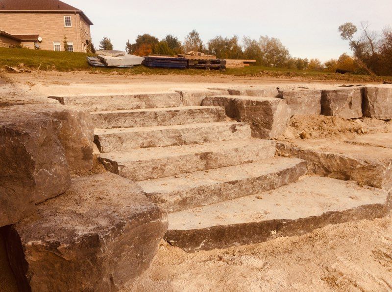 Stone steps leading down to a sandy area, with a house and trees in the background.