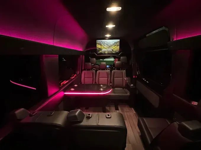 Interior of a luxury van with pink lighting, leather seats, a TV, and a table.