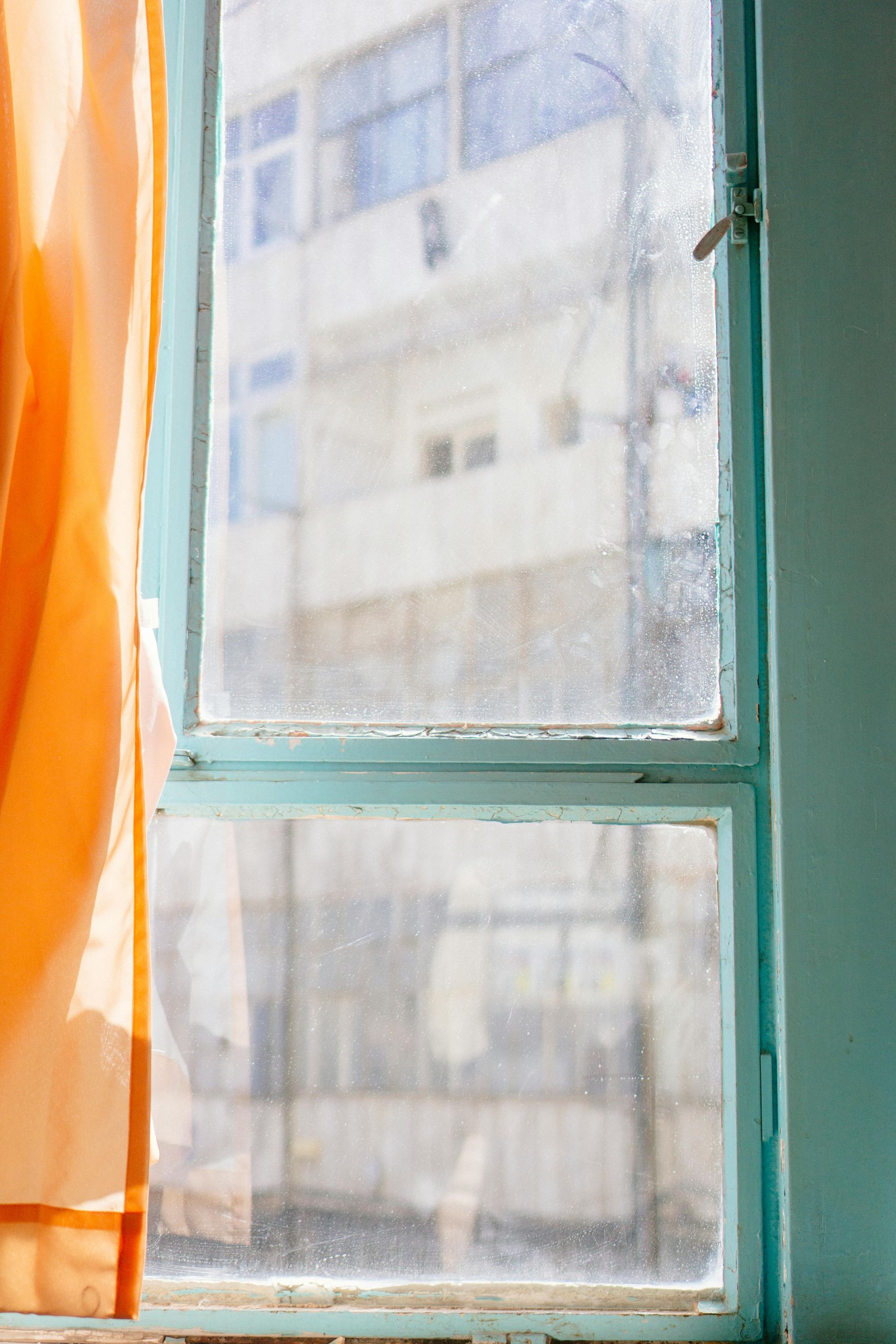 Blue-framed, dusty window with an orange curtain; blurry view of a building outside.