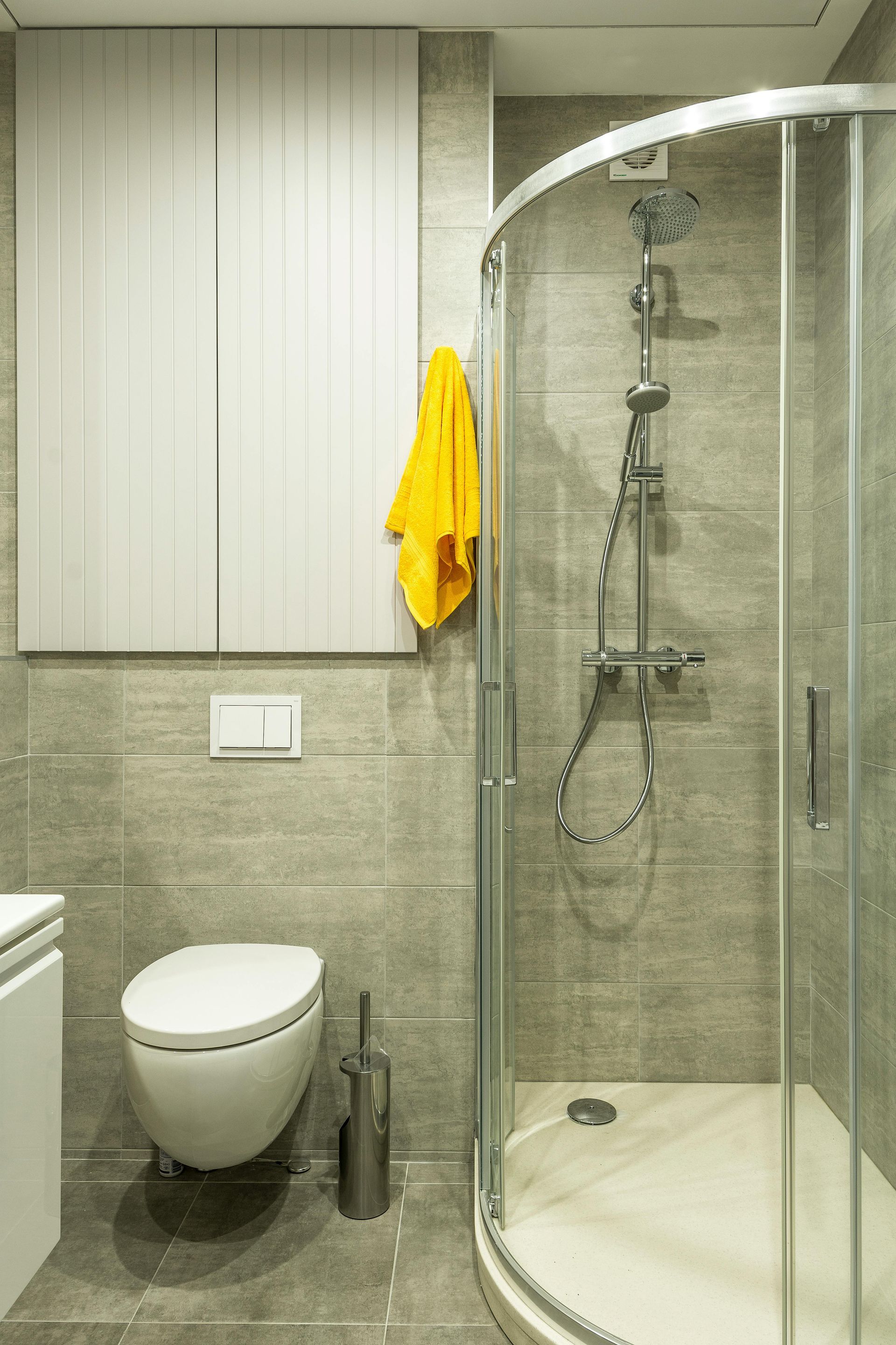 Small modern bathroom with gray tile, shower, toilet, and yellow towel.
