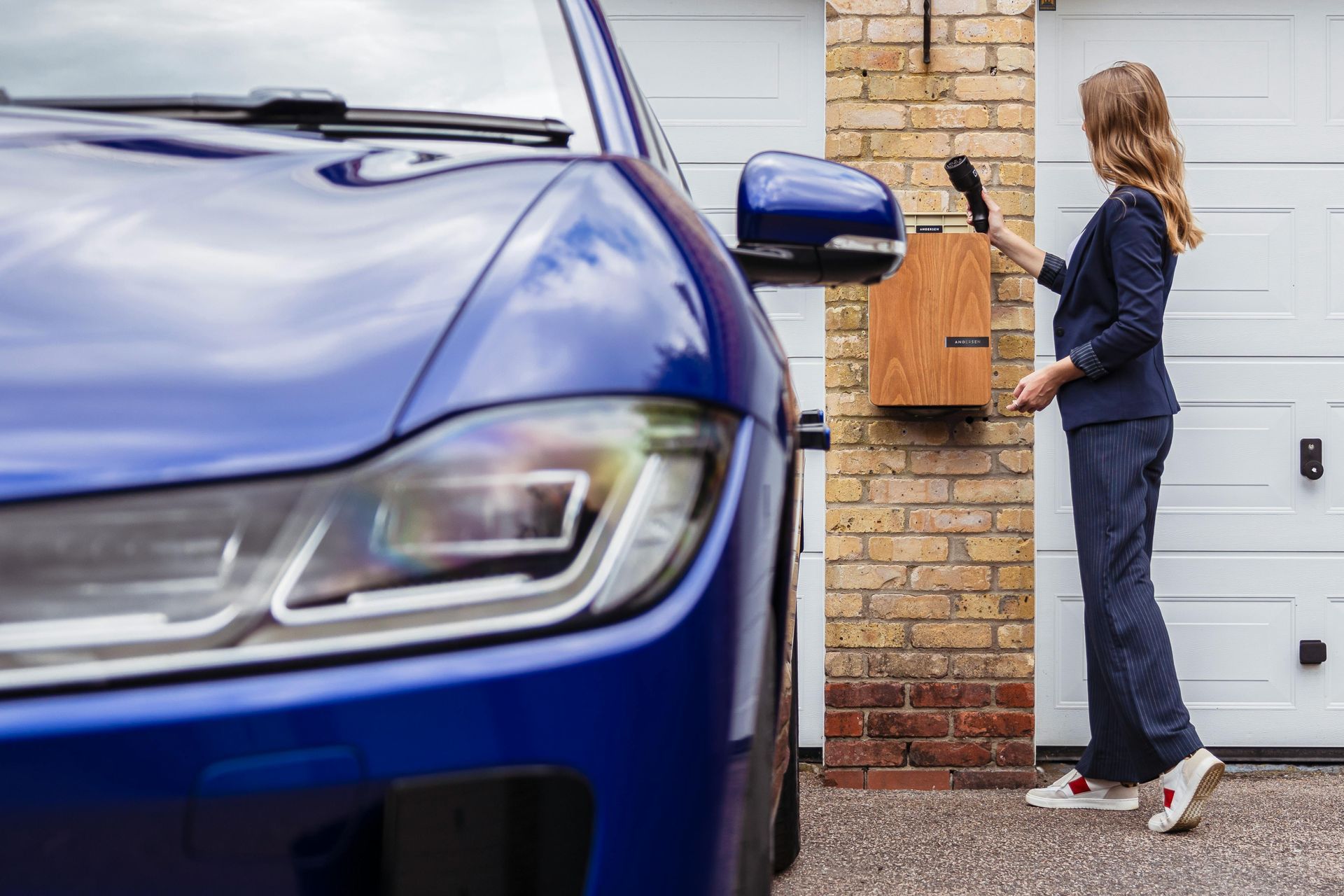 A woman in a blue suit checks a wooden mailbox next to a blue car and a brick garage.