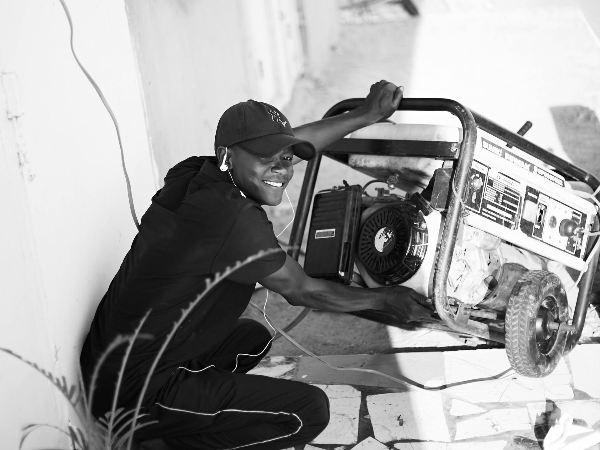 Man in cap, squats near a generator, smiles while holding it near a building.