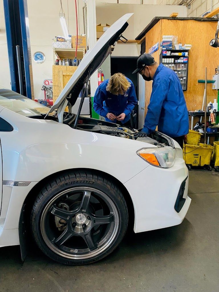 Two mechanics in blue uniforms work on a white car with an open hood in a garage. | Nutech Auto Repair