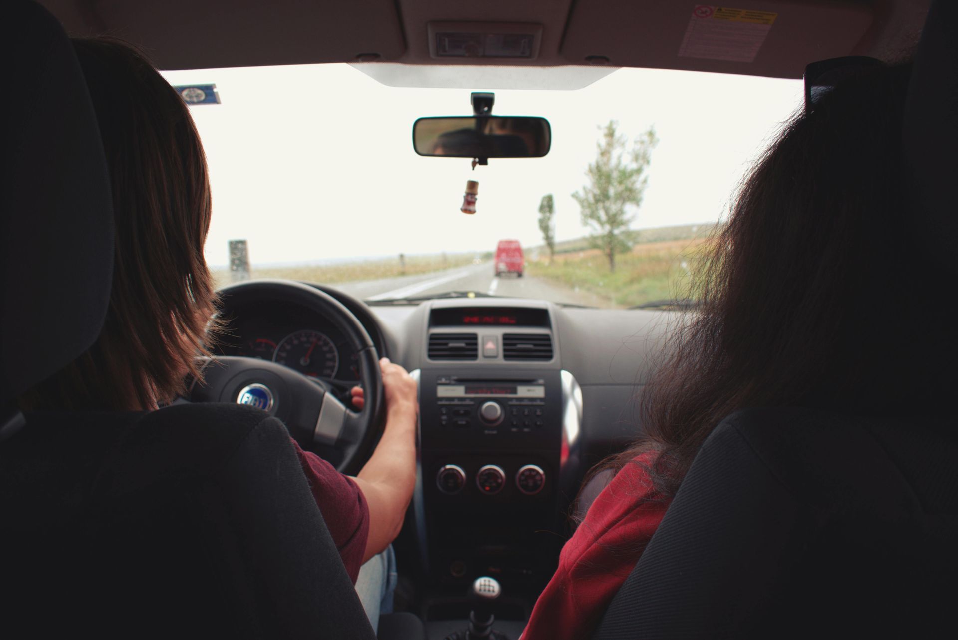 View from inside a car: a person driving on a road, a passenger, gray sky, and distant red car.