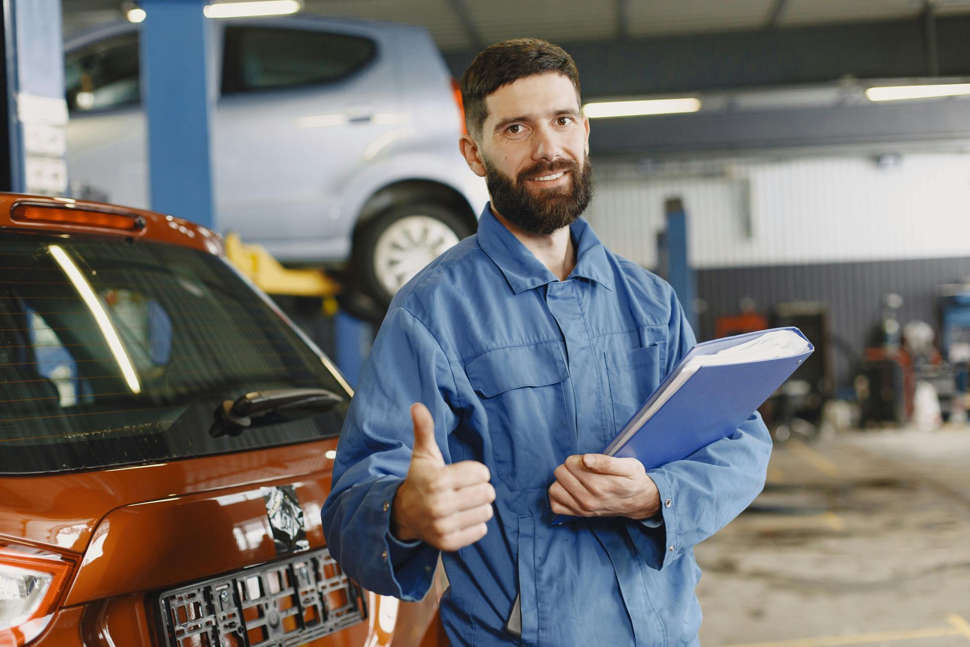 Mechanic in blue overalls with a beard gives a thumbs up, holding a clipboard in an auto repair shop next to a red car.
