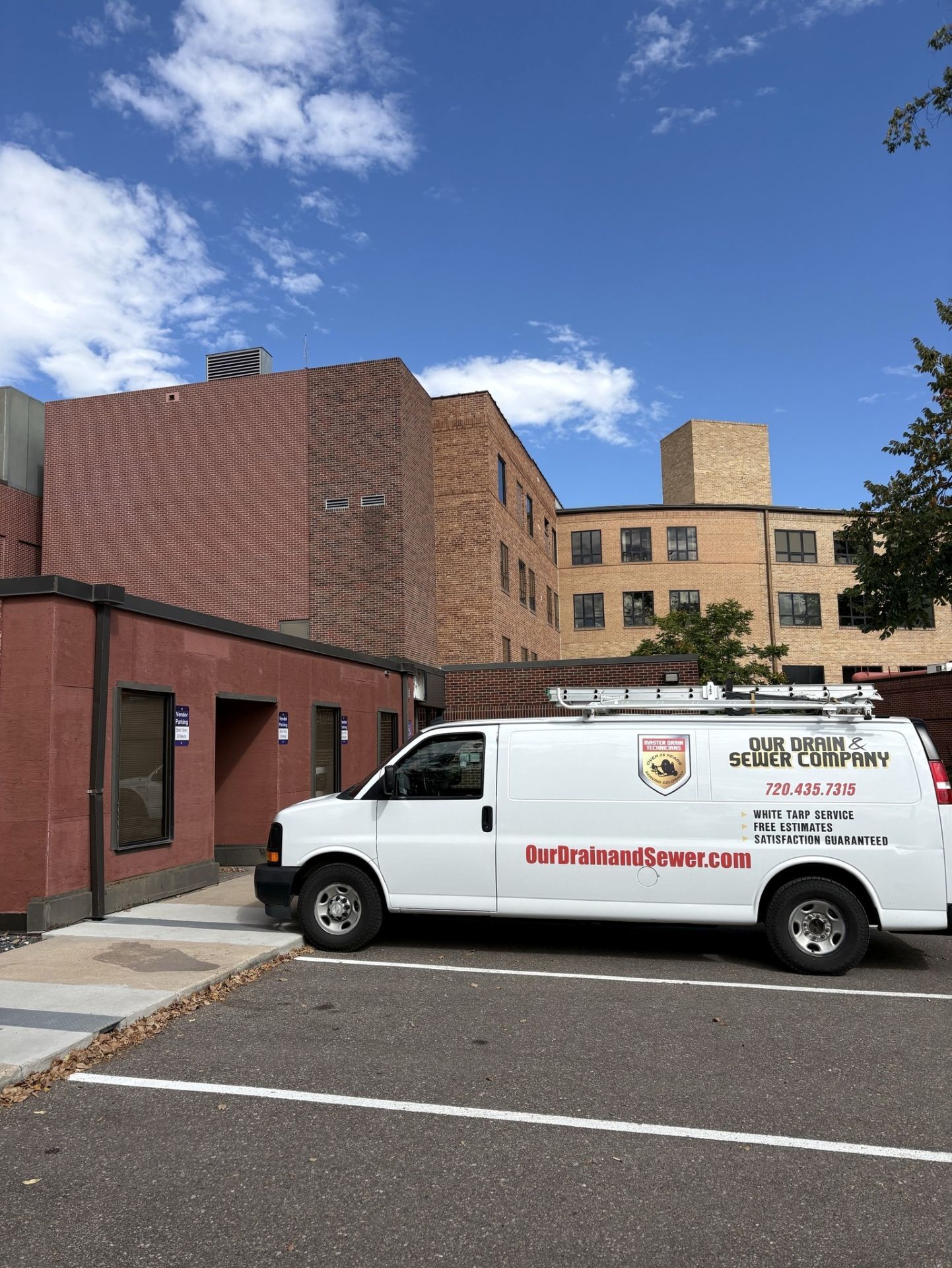A white service van parked in a paved lot next to a large red and tan brick building under a sunny, blue sky.