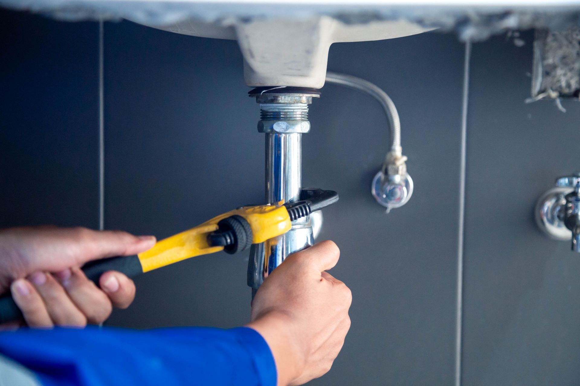 Hands wearing a blue uniform use a yellow pipe wrench to tighten a chrome drain pipe under a sink.