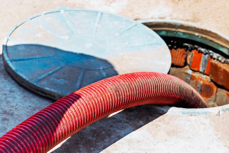 A thick, ribbed red hose inserted into an open manhole on a concrete surface.